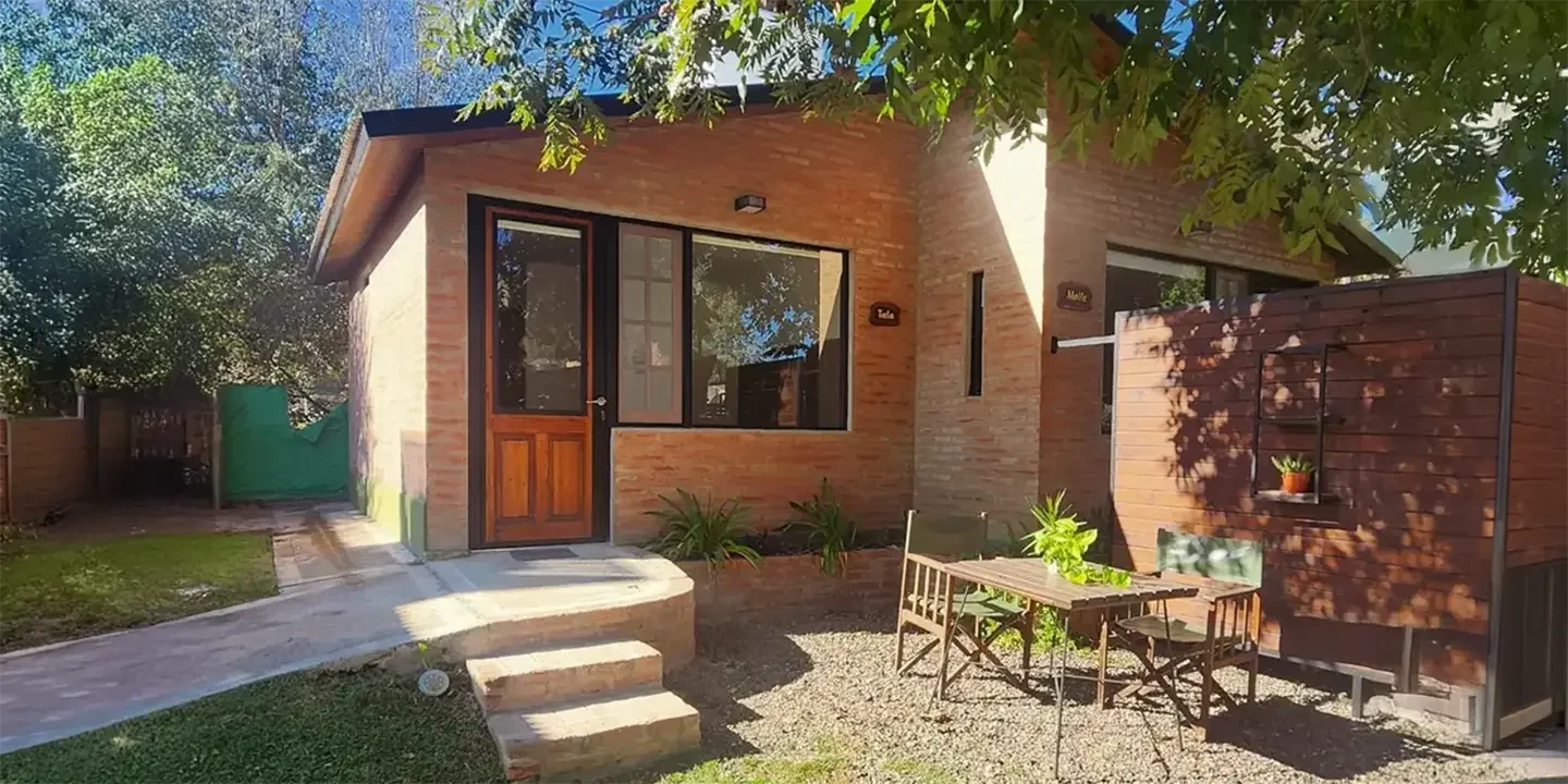 Vista exterior de una cabaña de ladrillo con entrada independiente, patio con mesa y sillas, rodeada de árboles y vegetación, ideal para descansar en un entorno natural.